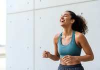 Young woman of mixed ethnicity wearing teal sports bra jogging indoors with bright natural light, expressing joy and fitness motivation for workout and exercise routines.