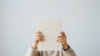 Person with medium skin tone holding open blank book in front of face, wearing light sweater. Suitable for education, learning, reading, knowledge, and publishing concepts.
