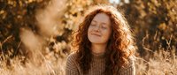 Young Caucasian woman with curly red hair laughing joyfully in golden autumn sunlight wearing cozy knit sweater for lifestyle and wellness content.