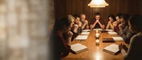 Business team praying together before meeting in warm conference room with documents and coffee cups on wooden table under pendant lighting.