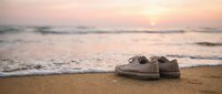 Pair of worn canvas shoes abandoned on sandy beach at sunrise with gentle ocean waves rolling in during golden hour.