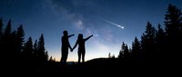 Silhouettes of two people standing on a hill pointing at a meteor streaking across the starry night sky surrounded by evergreen forest trees.