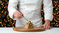 Professional chef in white uniform plating gourmet dessert on wooden board against colorful bokeh background for culinary presentation and fine dining.