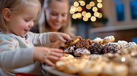 Caucasian mother and daughter baking Christmas cookies together, little girl reaching for chocolate treats on kitchen counter with festive bokeh lights.