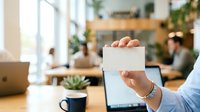 Businessman in light blue shirt holding blank white card in modern office workspace with colleagues working at wooden desk, laptop, potted plant, and natural window light.