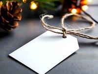 Blank white gift tag tied with rustic twine on dark background with pine cone and glowing holiday lights creating festive Christmas atmosphere.