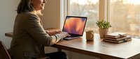 Middle-aged woman with light skin tone working on laptop at wooden desk by window with natural light, coffee cup, plant, and books nearby.