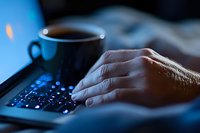 Male hand typing on laptop keyboard in dark room with coffee cup, illuminated by blue screen light creating moody atmosphere for remote work concept.
