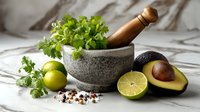 Fresh ingredients for guacamole preparation with stone mortar, wooden pestle, avocado, lime, cilantro, and spices on marble surface.