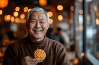 Cheerful senior Asian man holding traditional mooncake with blurred festive lights background, celebrating Mid-Autumn Festival in brown sweater.