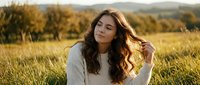 Young brunette woman with curly hair in cream sweater sitting in golden autumn field during sunset with mountains in background for lifestyle content.