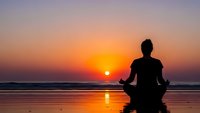 Woman practicing yoga meditation in lotus pose on beach at sunset with golden sky reflection on water for wellness and mindfulness concepts.