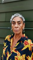 Senior woman with gray hair and Latin American ethnicity wearing a vibrant floral shirt with yellow and red flowers on navy background. Outdoor portrait against green wall backdrop.