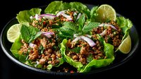 Fresh Thai-inspired minced meat lettuce wraps garnished with mint leaves, red onion, and lime wedges on dark plate against black background.