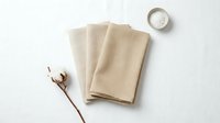 Folded beige linen napkins arranged on white surface with dried cotton branch and glass of water, flat lay composition for home decor and table setting design.