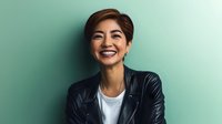 Young Asian woman with short hair wearing black leather jacket and white t-shirt laughing joyfully against mint green background, expressing genuine happiness and confidence.