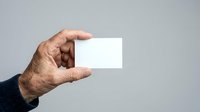 Close-up of mature male hand holding blank white business card against neutral gray background for corporate branding and professional networking purposes.