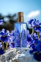 Blue glass perfume bottle with sparkling particles surrounded by purple iris flowers on white stone, soft bokeh background creates luxurious cosmetic atmosphere.