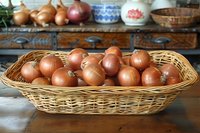 Fresh brown onions in woven wicker basket on rustic wooden table with vintage kitchen decor in background, natural food photography for cooking and farm markets.