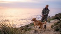 Middle-aged man with brown hair and beard, wearing casual outdoor clothing, walking dog on leash along coastal beach path during golden hour sunset with ocean and grass visible.