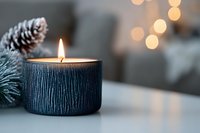 Glowing candle in textured black holder with pine cone and soft bokeh lights creating cozy winter atmosphere for holiday season and home decor.