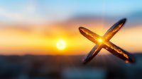 Wooden cross silhouette against vibrant orange sunset sky with glowing sun creating spiritual atmosphere. Symbolic religious photography for faith concepts.