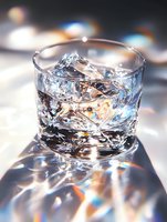 Crystal clear glass with ice cubes and light reflections creating rainbow prism effects, close-up macro photography on white surface with soft bokeh background.