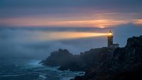 Lighthouse beacon shining over rocky coastline at dramatic sunset with golden sky and calm ocean waters creating peaceful maritime scene.