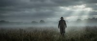 Lone figure walking through misty field at dawn with dramatic cloudy sky and fog rolling over rural landscape creating moody atmospheric scene.