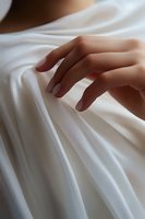 Elegant female hand gently touching soft white silk fabric with flowing folds and creases, creating sensual texture and light shadows for luxury fashion and beauty concepts.