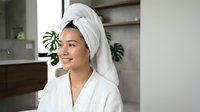Young Asian woman in white bathrobe and hair towel wrap smiling in modern bathroom, skincare and wellness routine concept.
