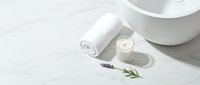 Rolled white towel, lit candle, and fresh lavender sprigs arranged on marble surface with ceramic bowl, spa and wellness styling.