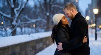 Romantic interracial couple embracing in snowy winter evening with warm street lights creating intimate atmosphere for Valentine's Day celebrations.