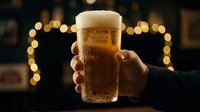 Hands holding cold beer glass with foam head against warm bokeh lights in dark bar setting, perfect for beverage advertising and hospitality marketing.