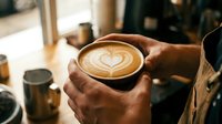 Hands holding cappuccino with heart latte art in cozy cafe setting. Morning coffee ritual, barista craftsmanship, warm atmosphere for hospitality business.