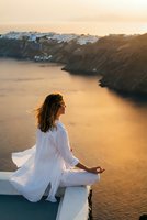 Young woman meditating in white flowing dress at sunset overlooking golden water and mountains. Peaceful mindfulness practice during golden hour retreat.