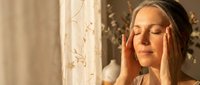 Young woman with closed eyes touching temples in peaceful meditation pose by sunny window with soft natural lighting for wellness and mindfulness concepts.