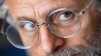 Close up portrait of mature bearded man wearing round glasses looking intently at camera with piercing blue eyes and serious expression.