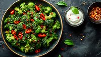 Fresh steamed broccoli florets with red chili peppers, sesame seeds and basil leaves served in dark bowl with yogurt sauce and spices on black background.