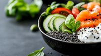 Fresh poke bowl with salmon, cucumber slices, black caviar, rice and basil leaves served in black ceramic bowl on dark background, healthy food.