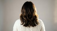 Young woman with wavy brown hair wearing white knit sweater viewed from behind in soft natural lighting for lifestyle and fashion concepts.