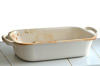 Empty white ceramic baking dish with handles showing signs of use and wear on rustic wooden surface, highlighting authentic kitchen character and cooking history.