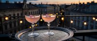 Two elegant pink cocktails on silver tray at rooftop terrace during sunset with city skyline and warm string lights creating romantic evening atmosphere.