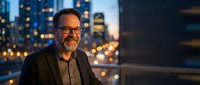 Confident mature businessman with glasses smiling at camera against blurred city lights background during evening hours for corporate portraits.
