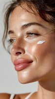 Close-up profile of woman with freckles and glowing skin applying facial cream on cheekbone against neutral background, showcasing natural beauty skincare routine.