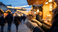 Hands in warm mittens holding hot chocolate with marshmallows at evening Christmas market with festive lights and crowd in background.