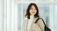 Young Asian businesswoman in beige coat smiling confidently in modern office building lobby with natural lighting for professional corporate content.