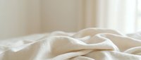 Soft beige linen fabric with gentle folds and wrinkles, creating texture and depth. Natural light illuminates the material, suitable for bedding, textile, and home decor projects.
