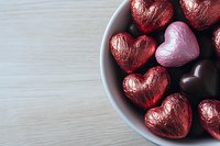 Heart shaped chocolate candies wrapped in red and pink foil arranged in white ceramic bowl on wooden table, romantic holiday celebration background with copy space.