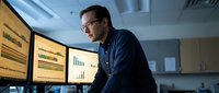 Professional Asian businessman analyzing financial data on multiple computer monitors in modern office environment late at night.